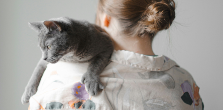 Vrouw met bruin haar in een knot en witte blouse met paarse en oranje bloemen heeft een grijze kat op haar schouder. Petrebels blog: top 10 vragen van kattenbazen over hun katten.